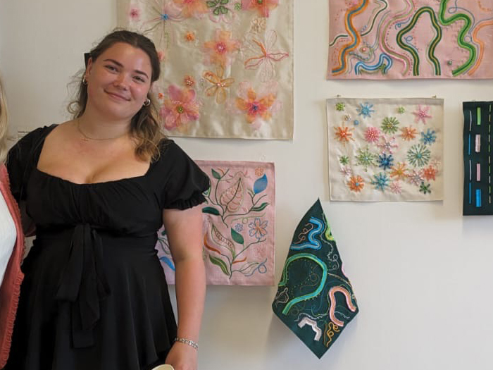 A person with long hair in a black dress standing in front of a wall with embroidered artwork