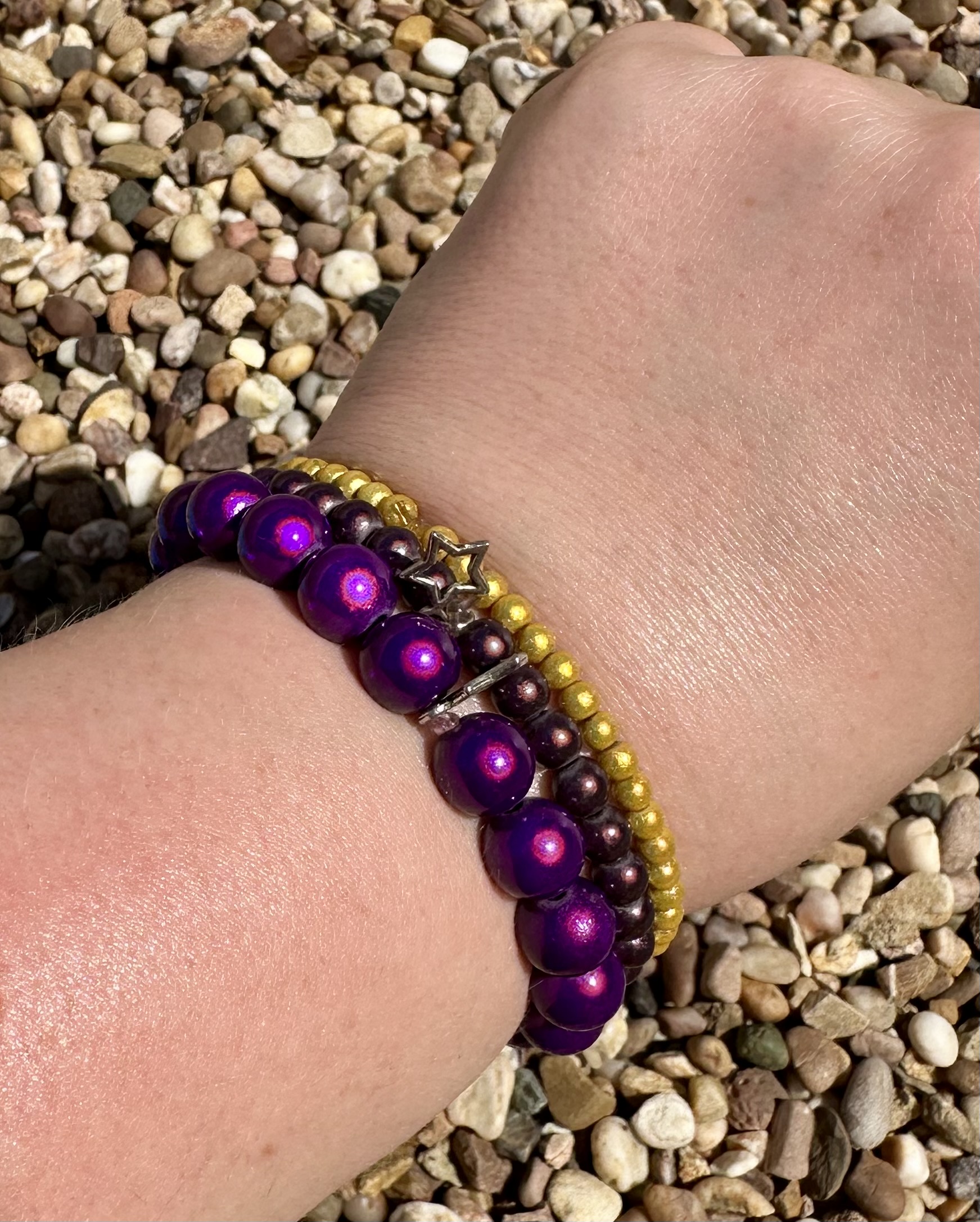 Hand wearing multiple purple and gold beaded bracelets including metallic purple beads and gold accent beads, photographed against a natural pebble background
