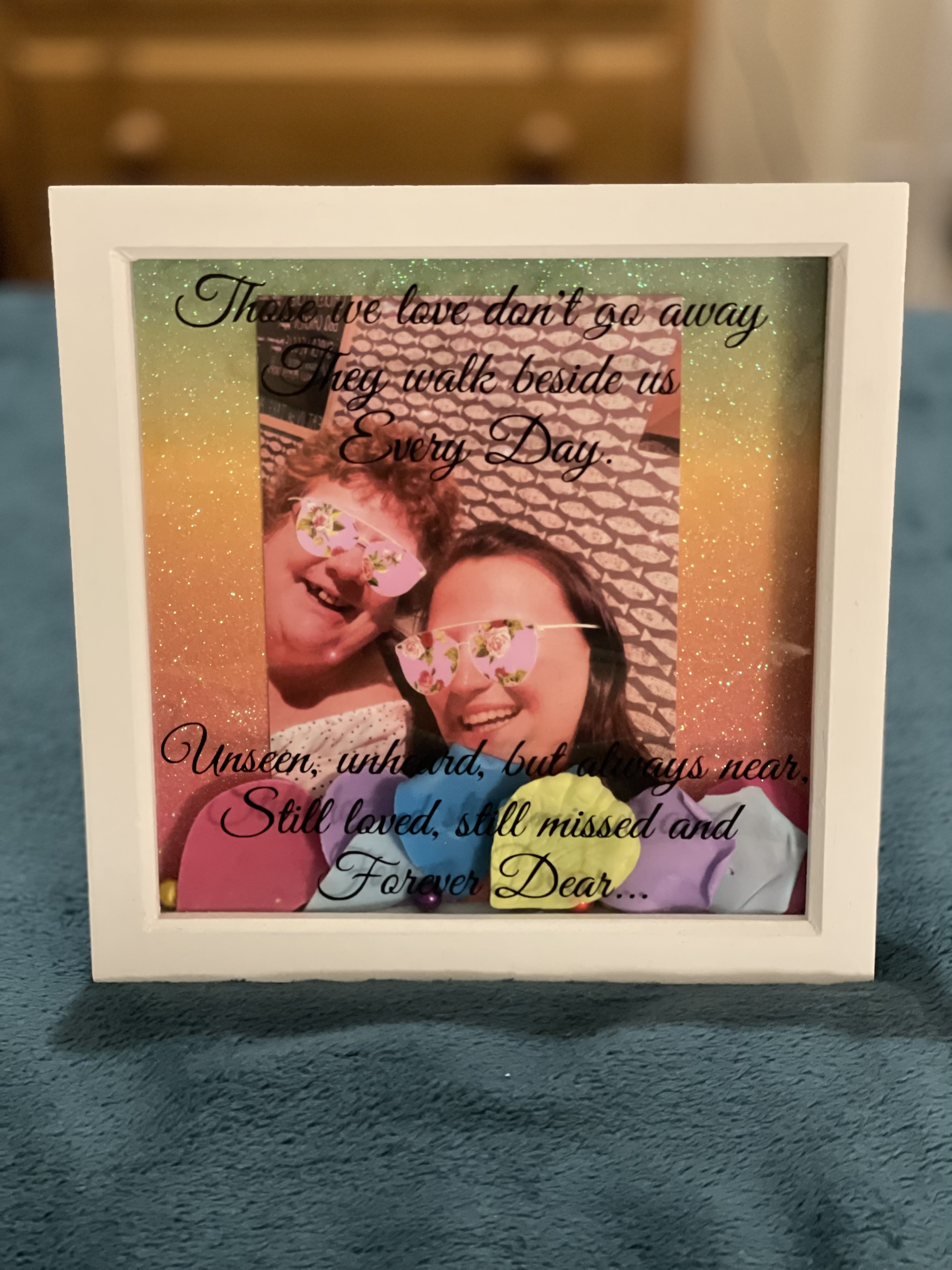 White framed memorial photo display with rainbow gradient background featuring two people wearing heart-shaped sunglasses, with text reading ‘Those we love don’t go away, they walk beside us every day’ and ‘Unseen, unheard, but always near, Still loved, still missed and Forever Dear