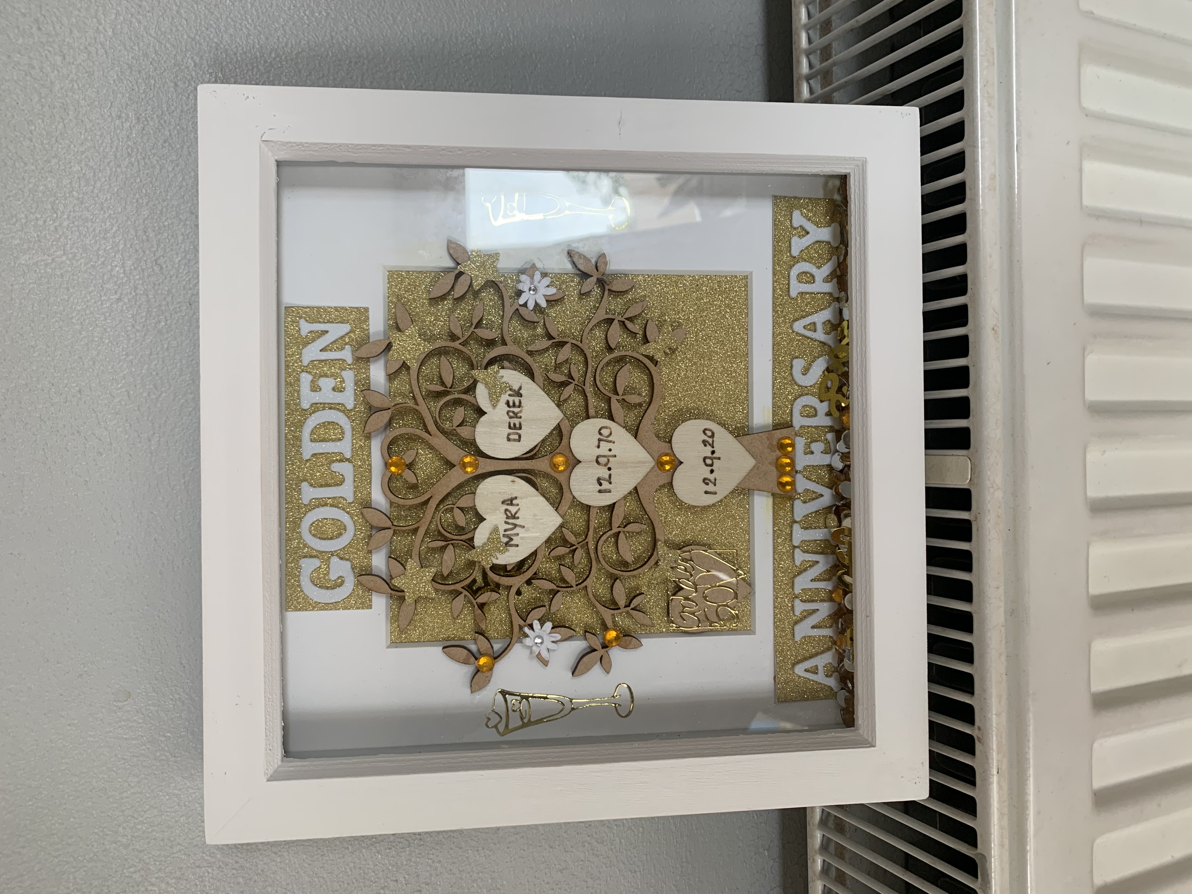 White shadow box frame displaying a golden anniversary design with decorative tree motif, hearts containing names ‘MYRA’ and ‘DEREK’ with dates ‘12.9.70’ and ‘12.9.20’, champagne glass decoration, and ‘GOLDEN ANNIVERSARY’ text with gold glitter background
