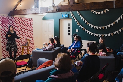a woman stands in front of a seated crowd. a creative cardiff banner and bunting hangs on the wall behind her